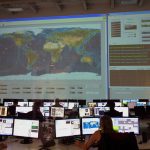 Profissionais analisam dados diante de computadores e de um grande mapa interativo em uma sala de controle.