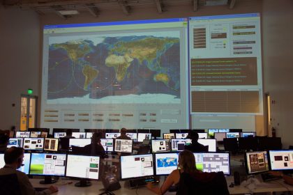 Profissionais analisam dados diante de computadores e de um grande mapa interativo em uma sala de controle.