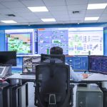 Operador acompanha sistemas em uma moderna sala de controle, diante de painéis e telas de monitoramento