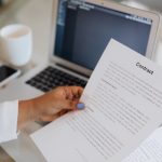Pessoa segurando um documento jurídico ao lado de um laptop em um ambiente de trabalho.